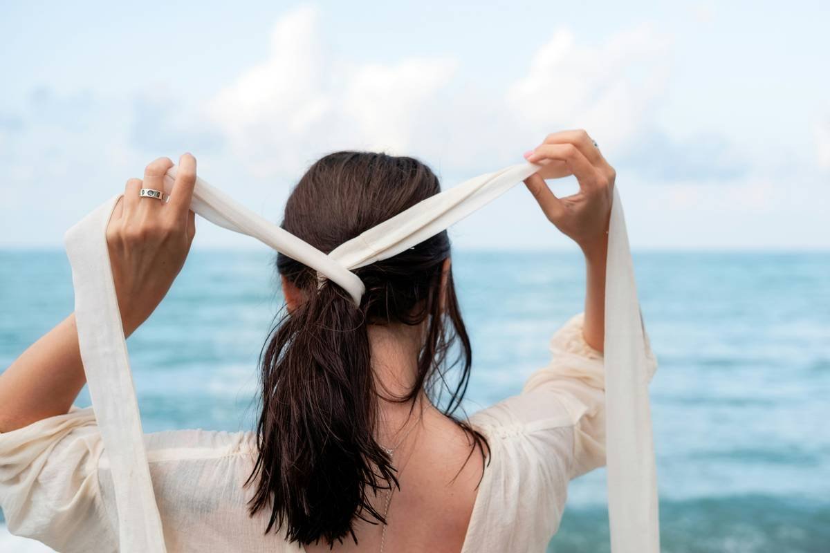 A girl in a wedding dress on the sea ties a ribbon in her hair