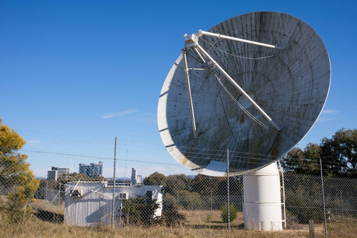Broken satellite dish against blue sky, inside a fence with highrise buildings in background.