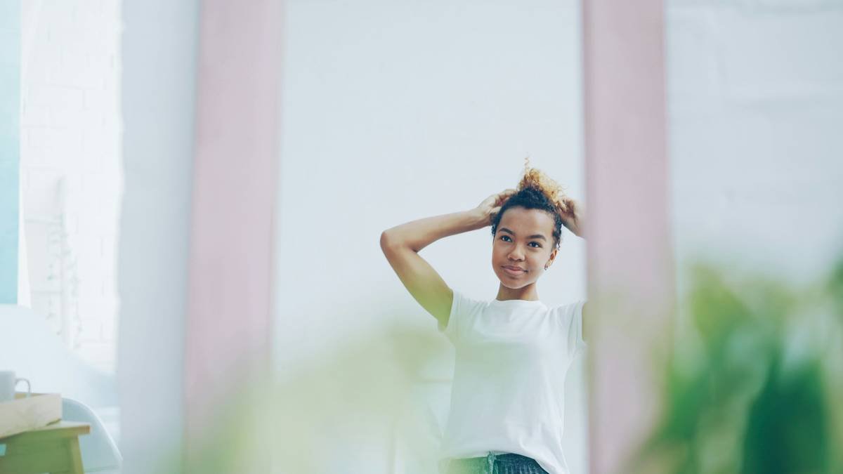 Pretty African American girl is fixing her curly hair looking in the mirror at home and smiling. Beautiful young people, youth lifestyle and interiors concept.